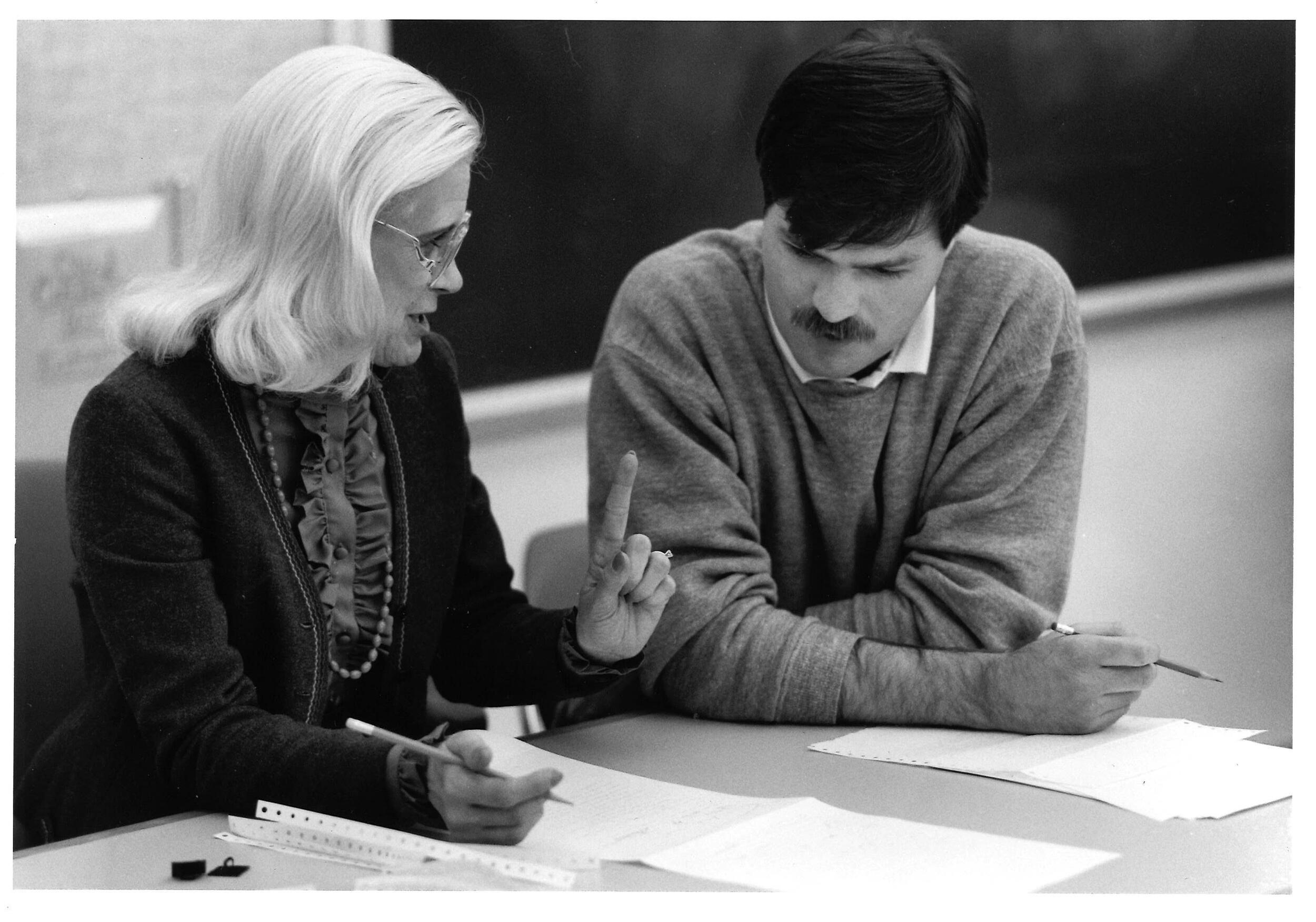 A photo of a man and a woman sitting at a table together. The woman is holding a pencil and is resting her hand on some papers that are in front of her. The woman is talking and the man is looking down at the papers in front of the woman. 
