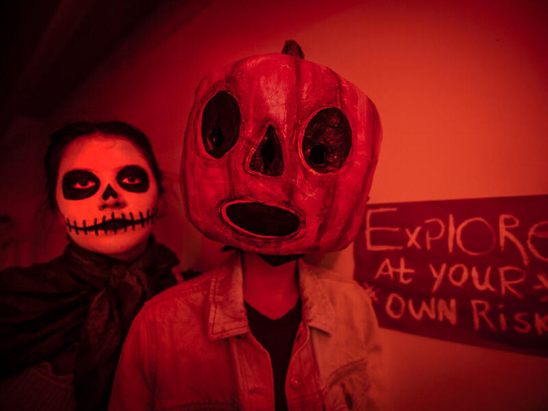 A person with skeleton makeup standing next to another person wearing a jack-o-lantern face mask. Next to them is a sign that says \"EXPLORE AT YOUR OWN RISK.\"