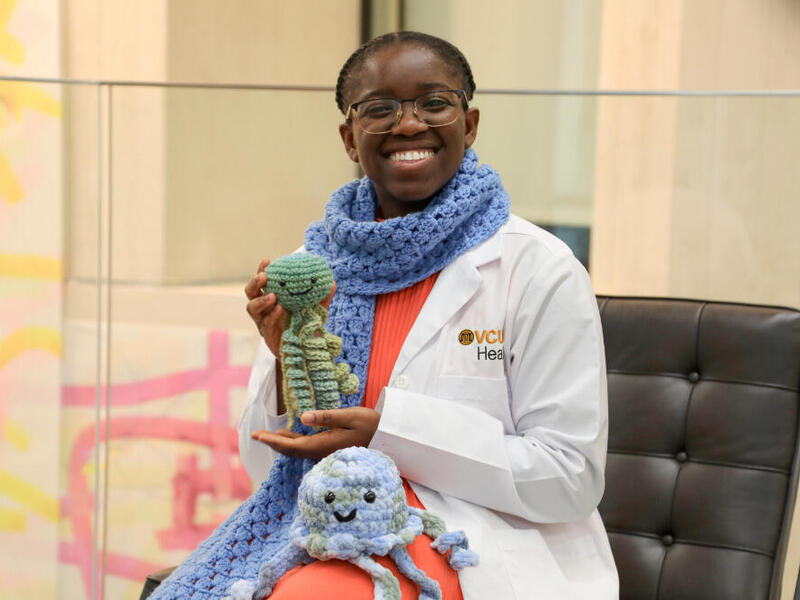 A photo of a woman sitting in a chair wearing a scarf and holding a knitted plush toy. There is a knitted jellyfish also sitting on her lap. 