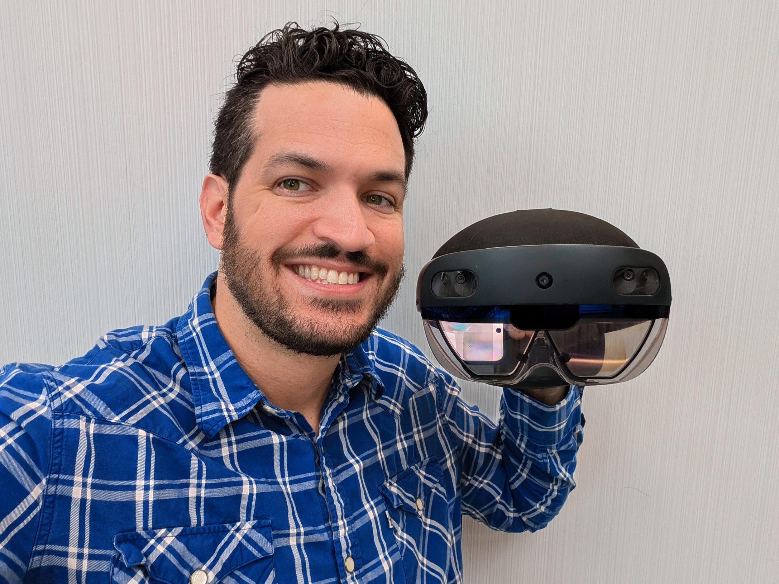 A photo of a man from the shoulders up. He is smiling and holding a VR headset in his right hand. 