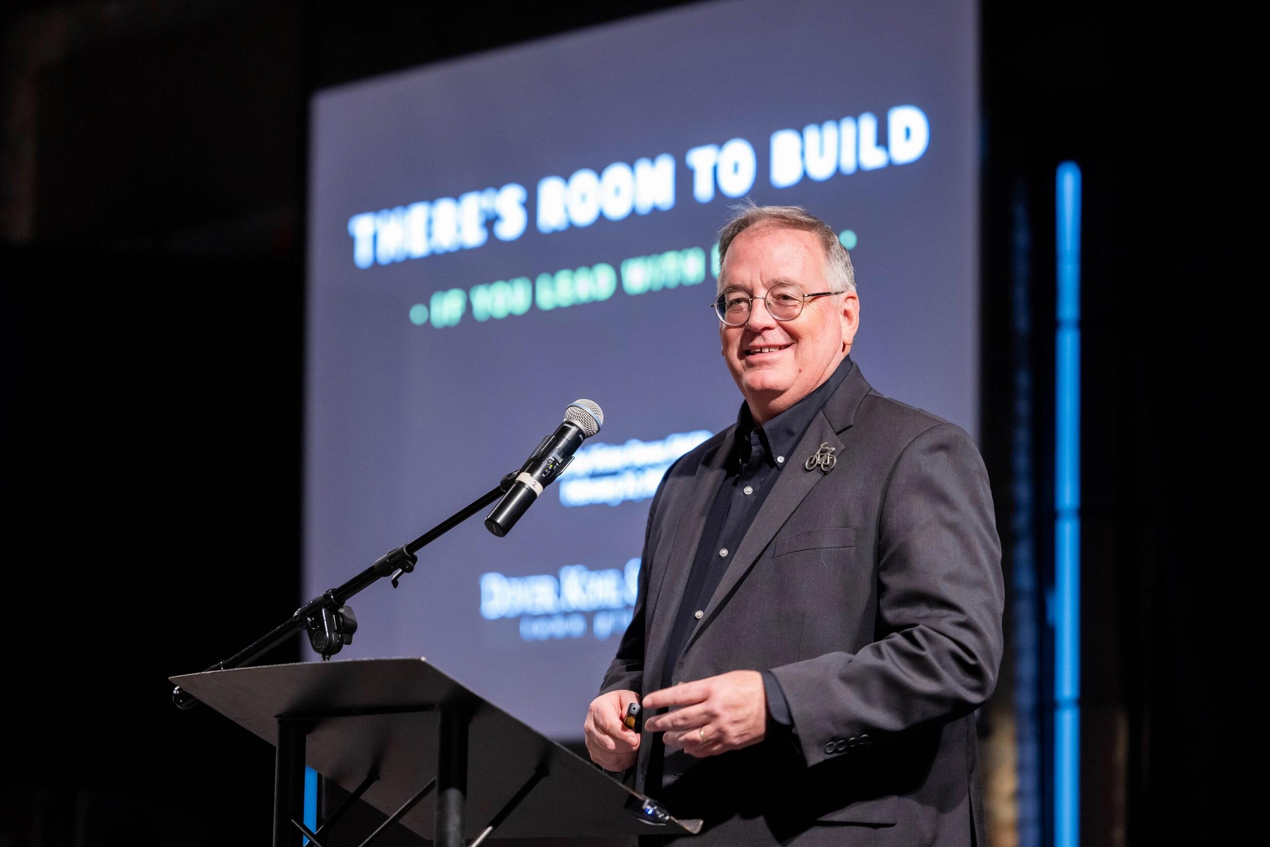 A photo of a man standing behind a podium with a micophone. 