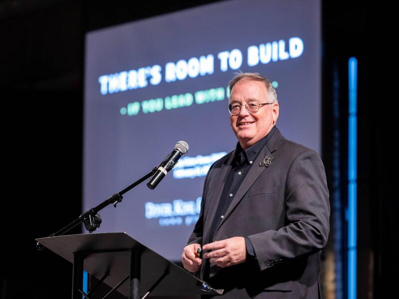 A photo of a man standing behind a podium with a micophone. 