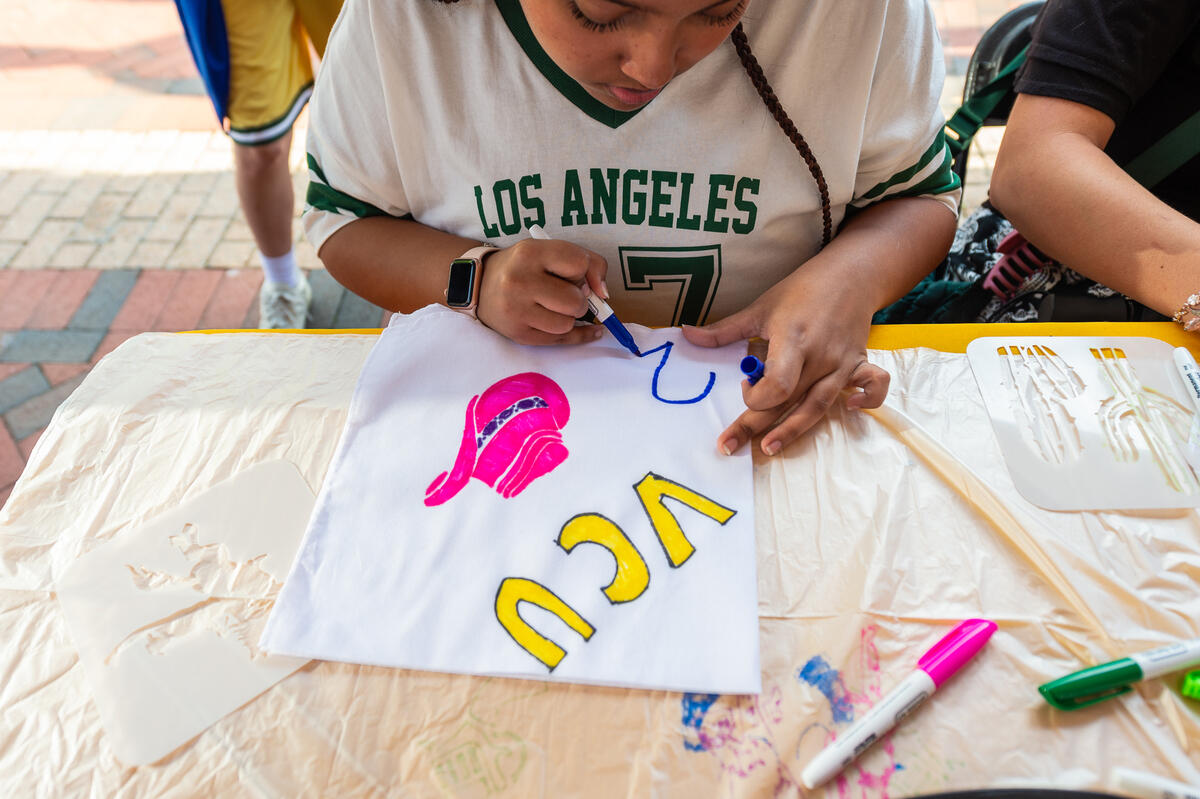 A photo of a student drwaing on a white square. The square says \"VCU,\" has an illustration of a pink cowboy hat, and the student is writing the number two. 