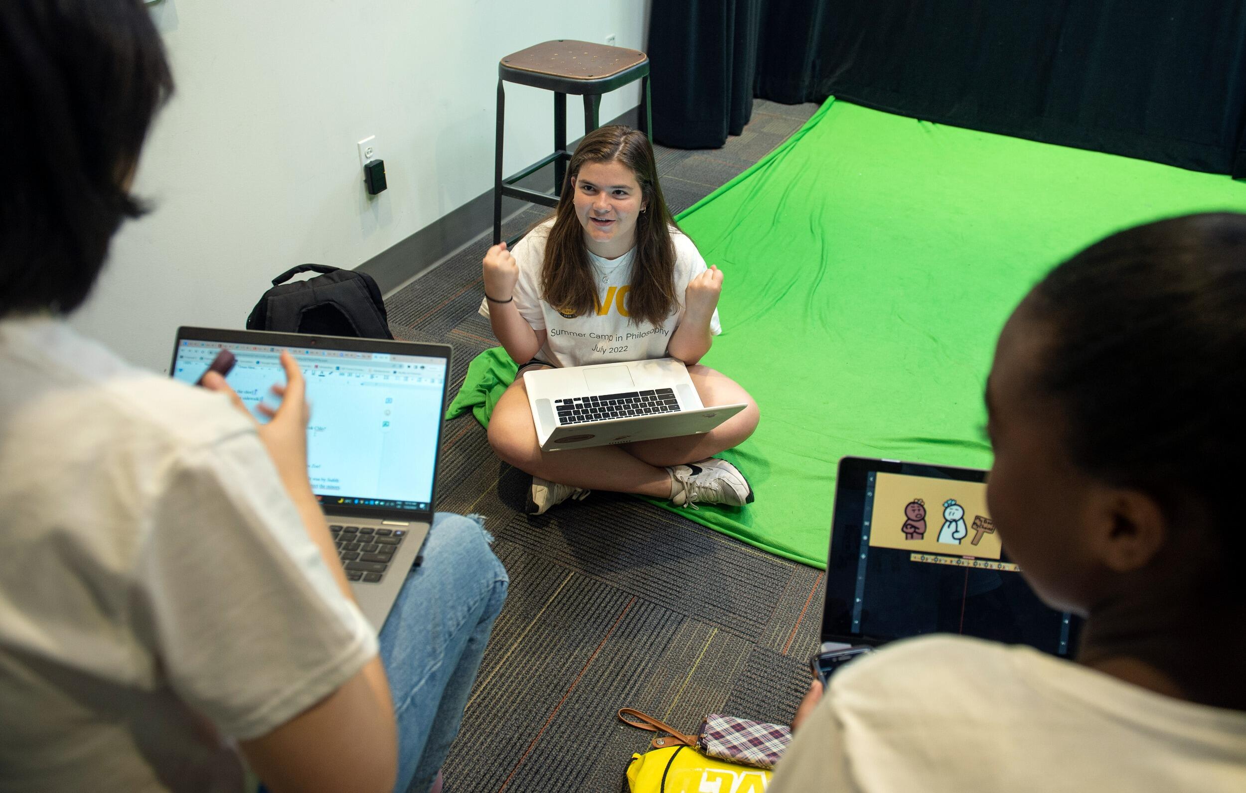 A high school student sits on the floor with a computer in her lap and speaks to two fellow students with computers who are facing her.