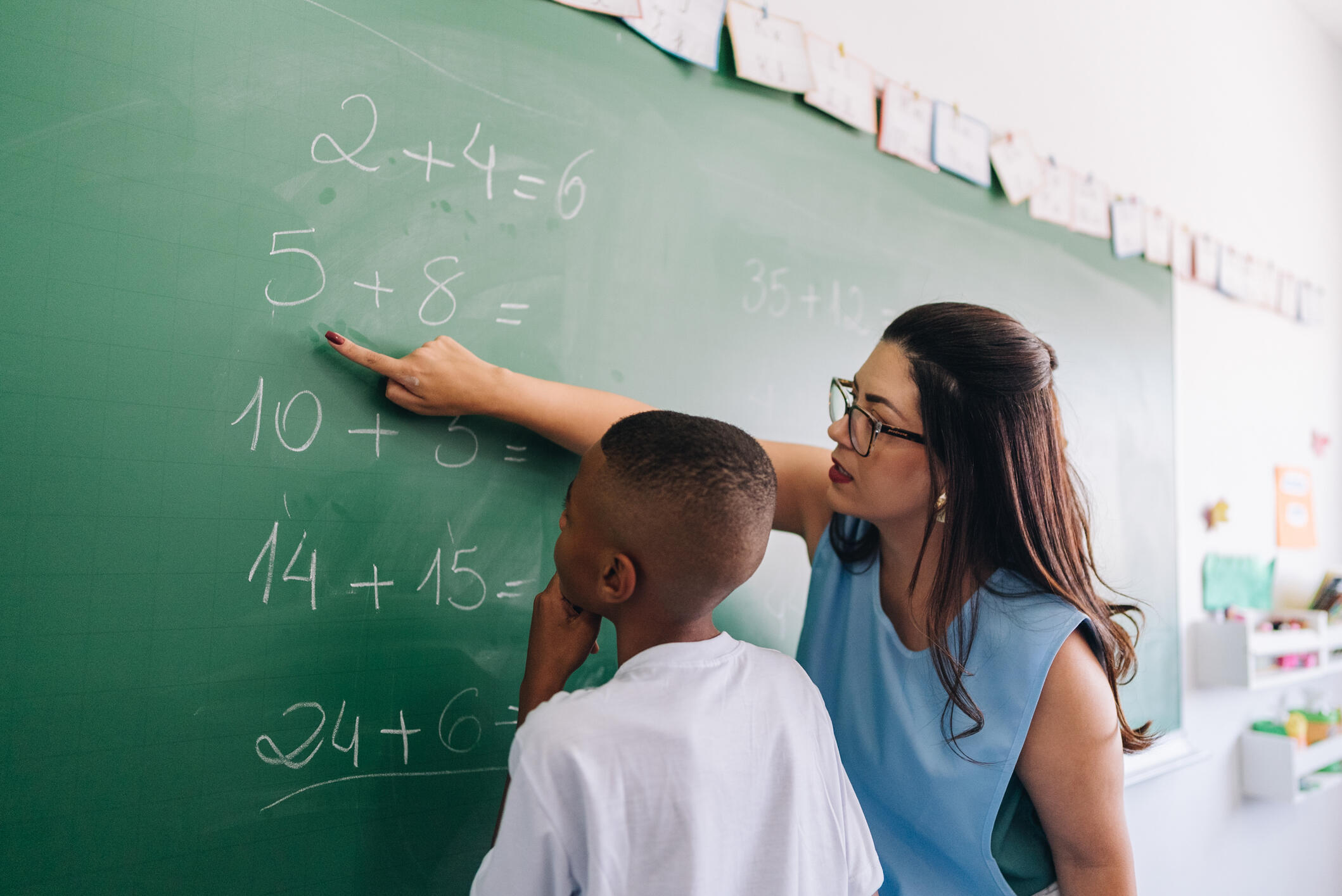 A photo of a woman pointing at a math problem on a chalk board. A boy is standing next to her looking at the problem. 