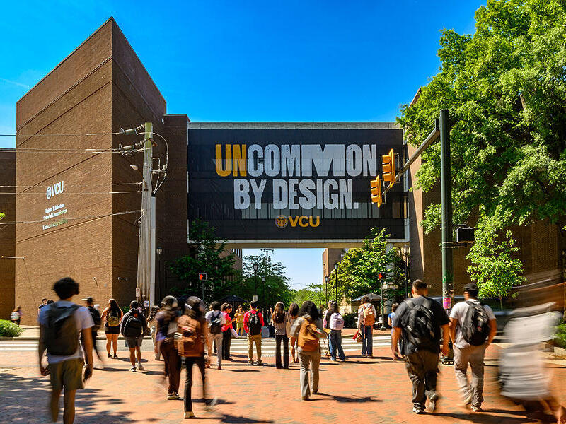 A photo of people walking on a sidewalk. On the building across the street is a large sign that says \"UNCOMMON BY DESIGN VCU\" 