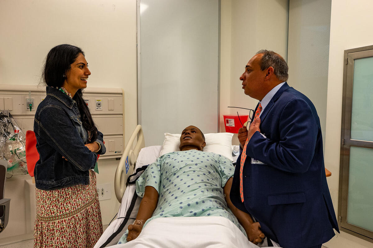 A photo of a man and women talking to each other on either side of a hospital bed with a dummy in it. 