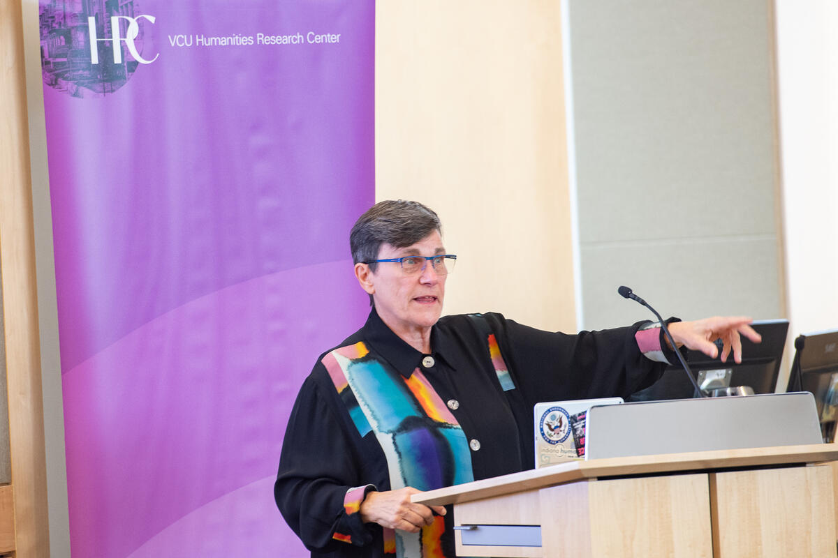 A photo of a person speaking behind a podium with a microphone on it. The person has their left arm extended in front of them. 