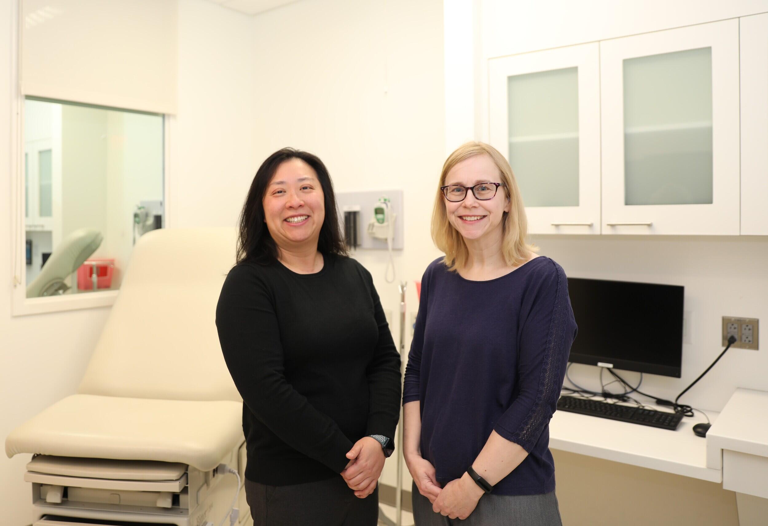 A photo of two women standing in a medical examination room. Behind them is a medical examination table and a countertop with a computer monitor and keyboard on it. 