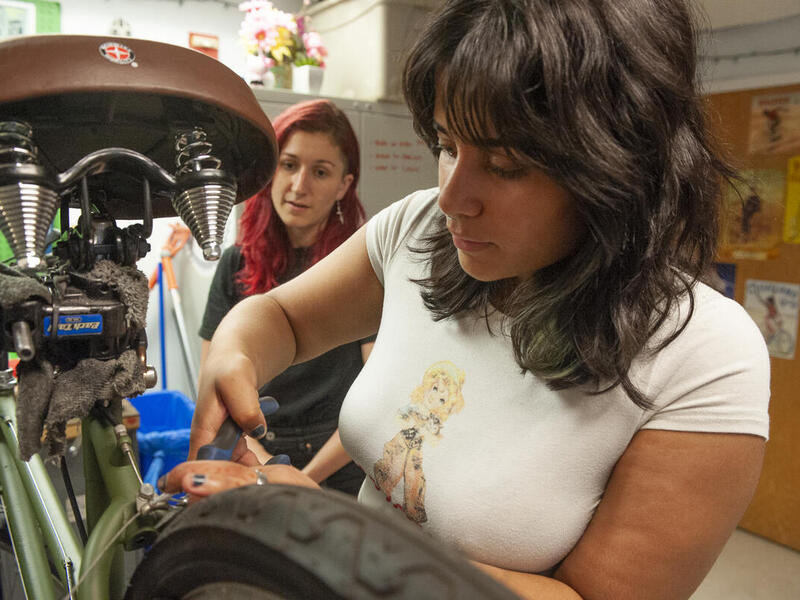 A woman working on a bike with another woman watching her. 