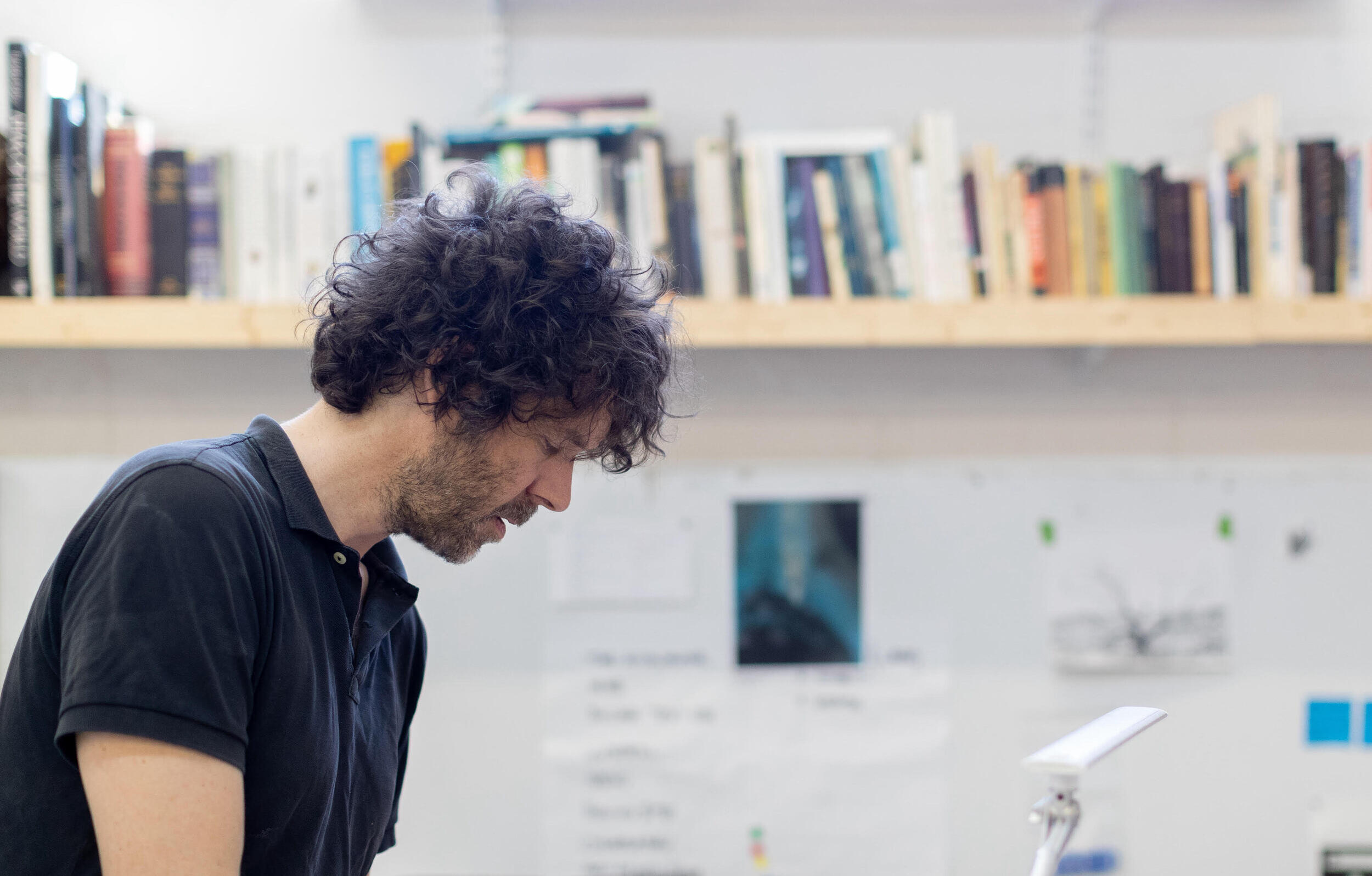 A photo of a man from the chest up. He is looking down at something not in the photo. Behind him is a shelf with books. 
