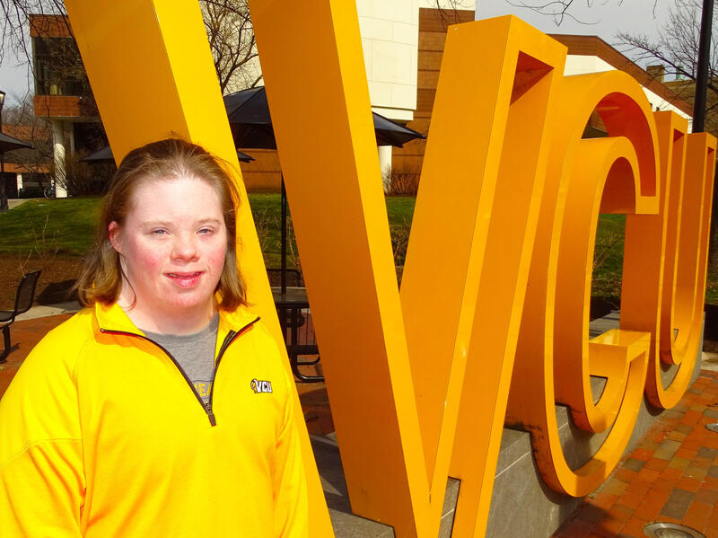 A photo of a woman standing next to giant yellow letters that spell out \"V C U\"