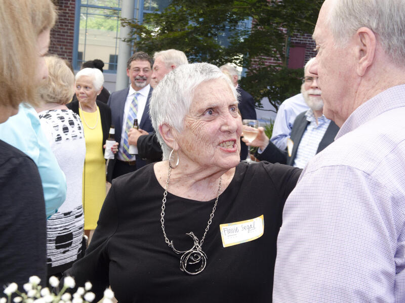 A photo of a woman talking to a man on her right. Around and behind them are several people who look like they are socializing with each other. 