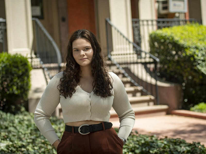 A photo of a woman standing outside with her hands in her pockets. 