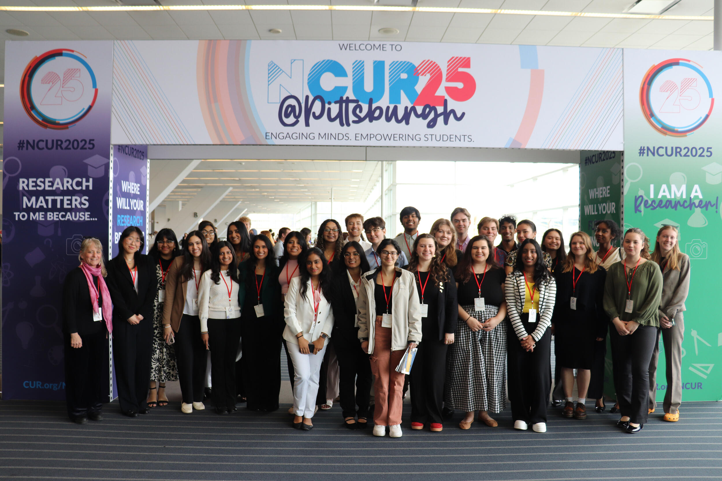 A group photo of 31 people standing under a banner that says \"NCUR25\" \"@Pittsburgh\"