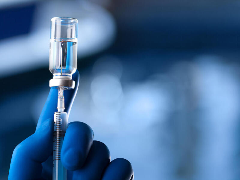 A glove-wearing hand holding a vial with an inserted hypodermic needle.