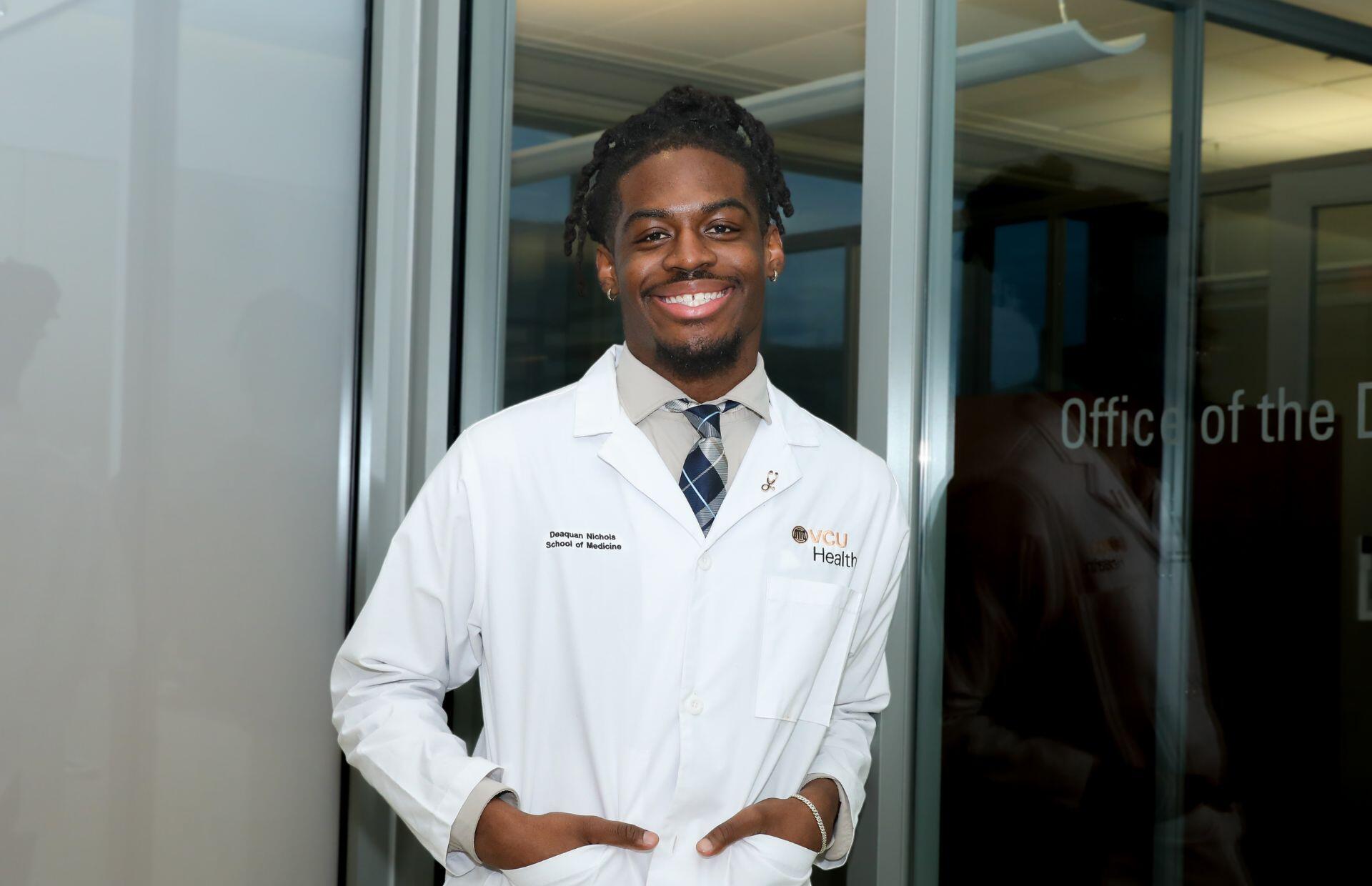 A photo of a man fromt he waist up. He is simling with his hands in his pockets and wearing a white lab coat. 