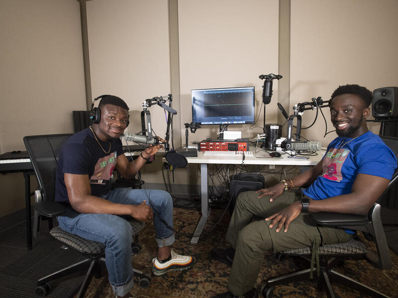 Roland Kusi and Atuahene Adu-Gyamfi sitting at a desk with podcasting equipment while wearing headphones
