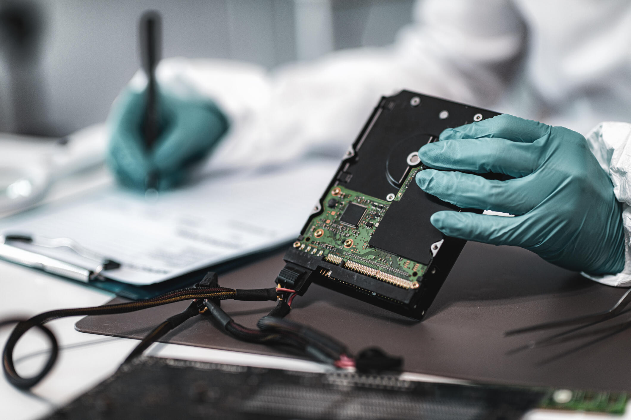 A photo of a person wearing blue latex gloves holding a circuit board in one hand and writing on a clip board with the other. 