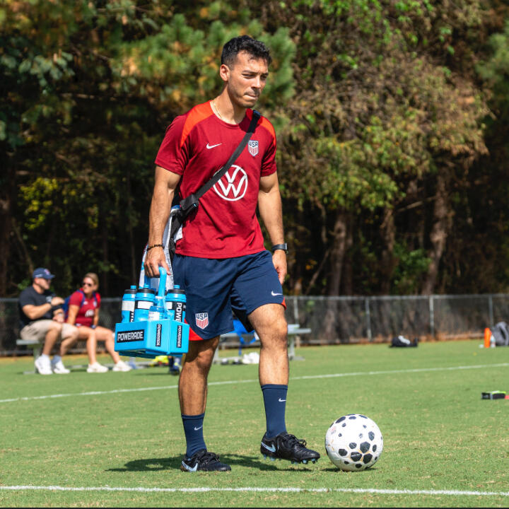 A man in a soccer uniform is standing on a field while holding a water bottle carrier and standing next to a ball. 