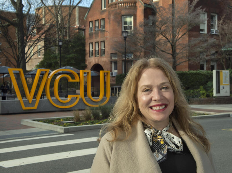 A photo of a woman from the shoulders up standing outside across the street from a sign that says \"VCU\" in yellow letters. 