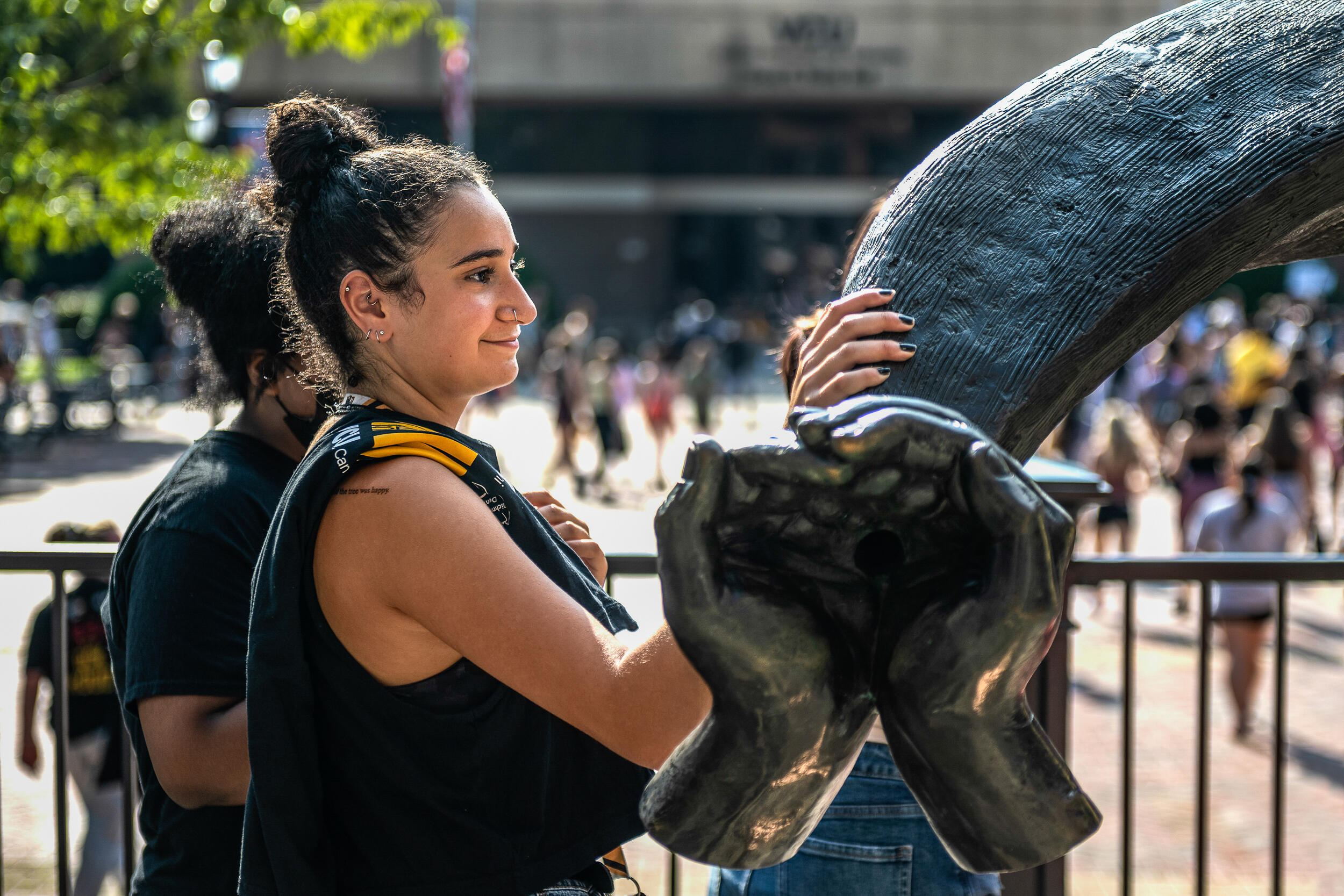 A student with hair in a bun and a New Student Orientation shirt over their shoulder rests a hand on a sculpture of ram horns in front of a plaza in the afternoon sun.
