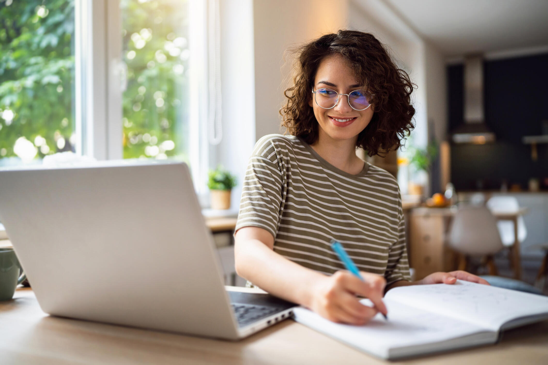 A woman in glasses writes in a notebook and looks at a laptop screen.