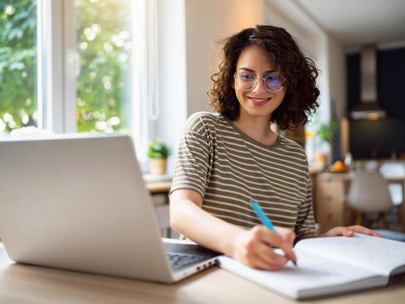 A woman in glasses writes in a notebook and looks at a laptop screen.