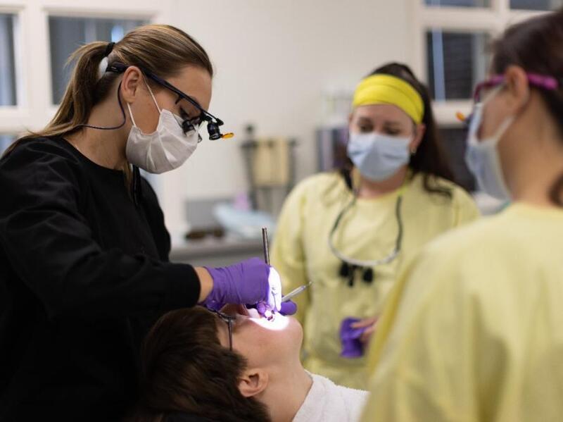 Dental students and a supervisor in surgical masks work with a patient.