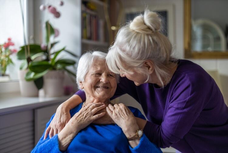 A photo of a woman wrapping her arms around an elderly woman with gray hair. Both women are smiling. 