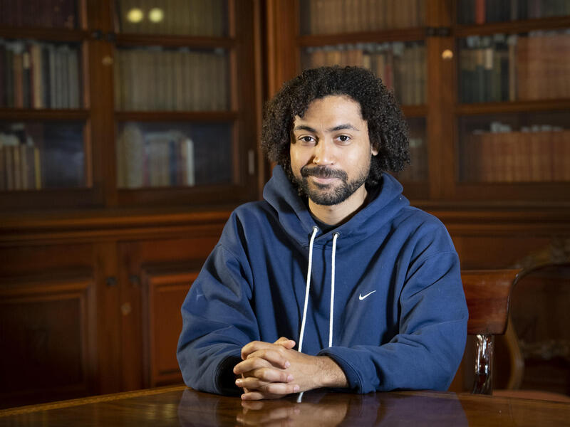 A photo of a man sitting at a table in front of bookcases. 