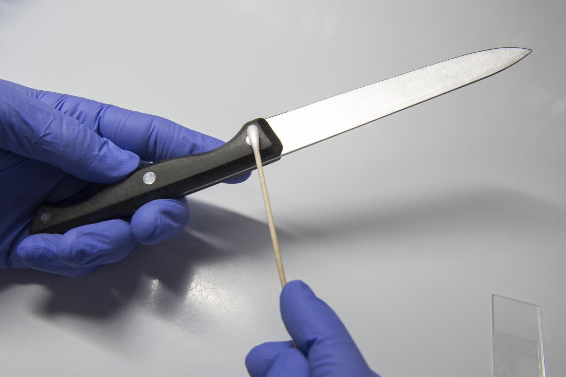 A person wearing blue gloves holds a kitchen knife and swabs it with a cotton swab.
