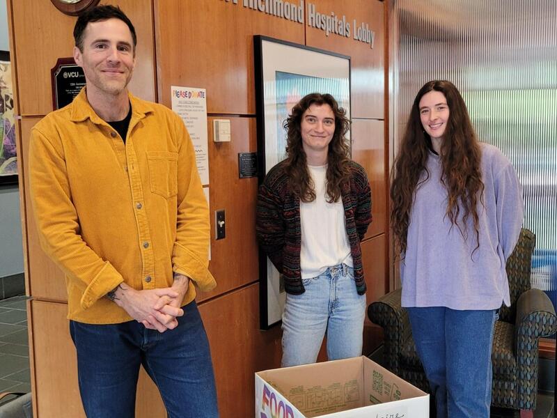 A photo of three people standing. On the left is a man and in the middle and right are two women. There is a white box in front of the group which says \"FOOD DRIVE\" and \"RVA\" in rainbow letters. 