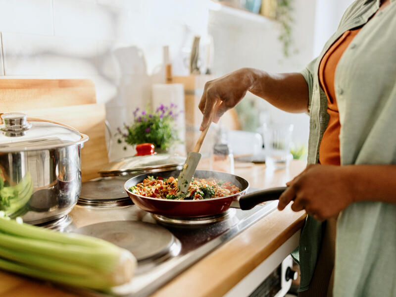 A photo of a woman from the neck down cooking food in a frying pan on a stove. 