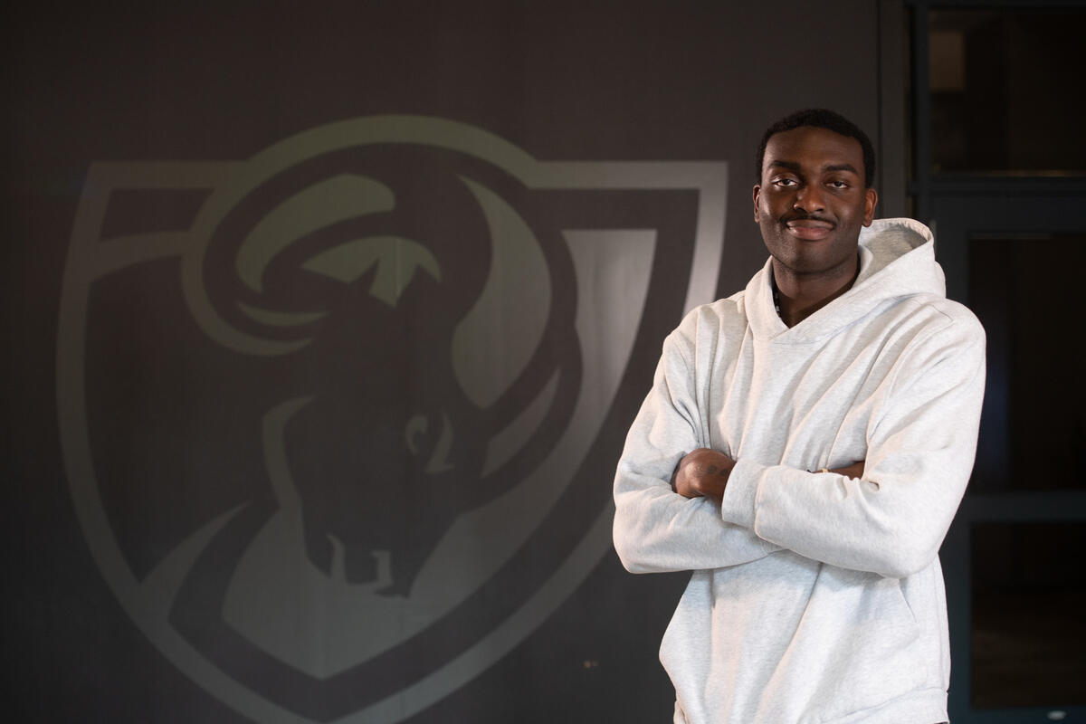 A man in a white hooded sweatshirt stands with his arms crossed in front of the VCU Rams logo.