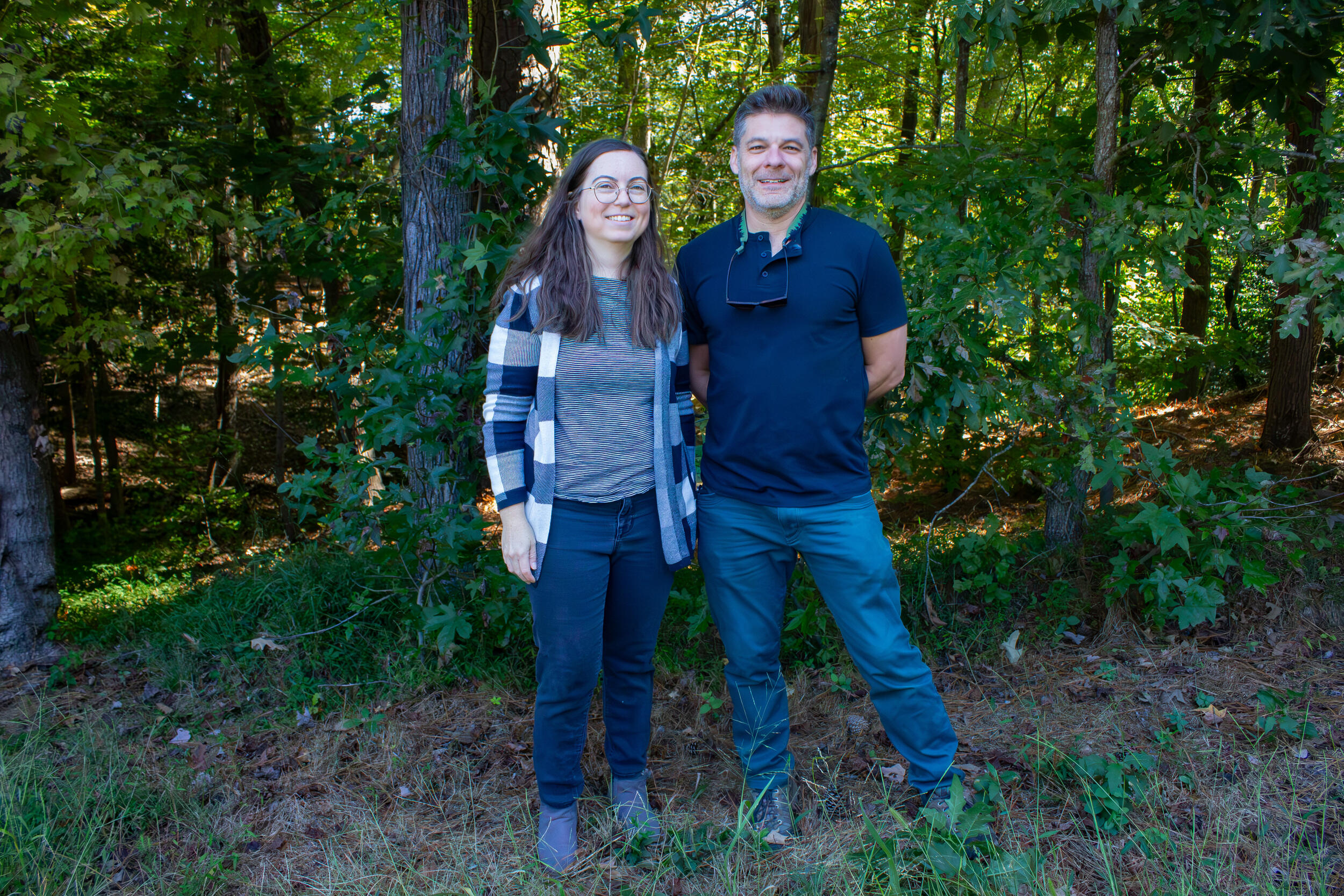 A photo of a man and woman standing at the edge of a forest and smiling. 