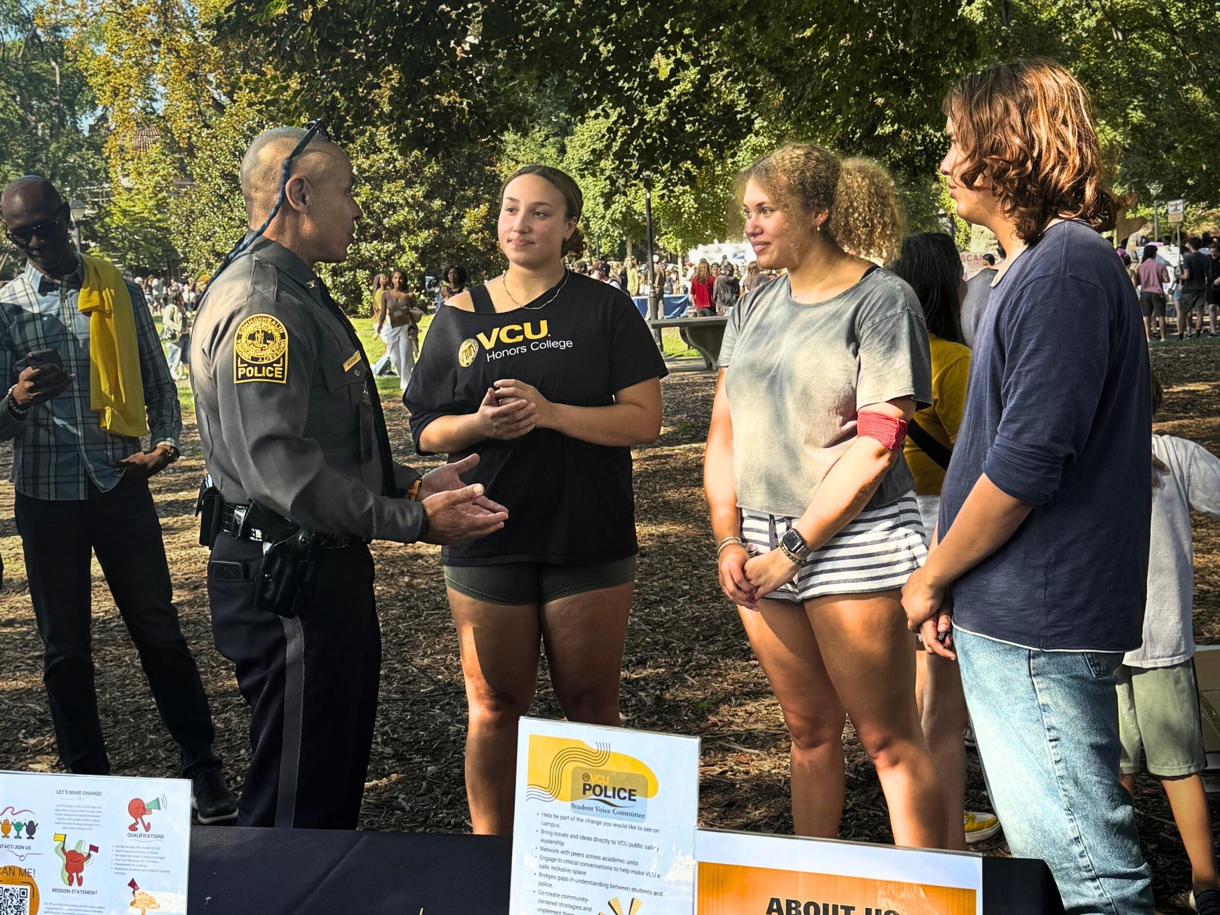 A photo of a police officer speaking with three students in a park. Behind them are other people milling about the park. 