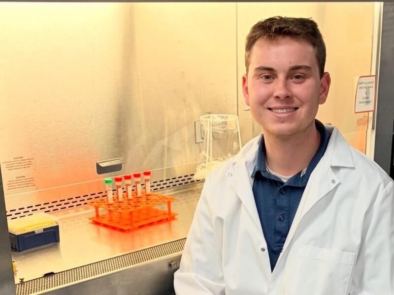 A phto of a man wearing a lab coat sitting in front of an open air vent with test tubes in it. 