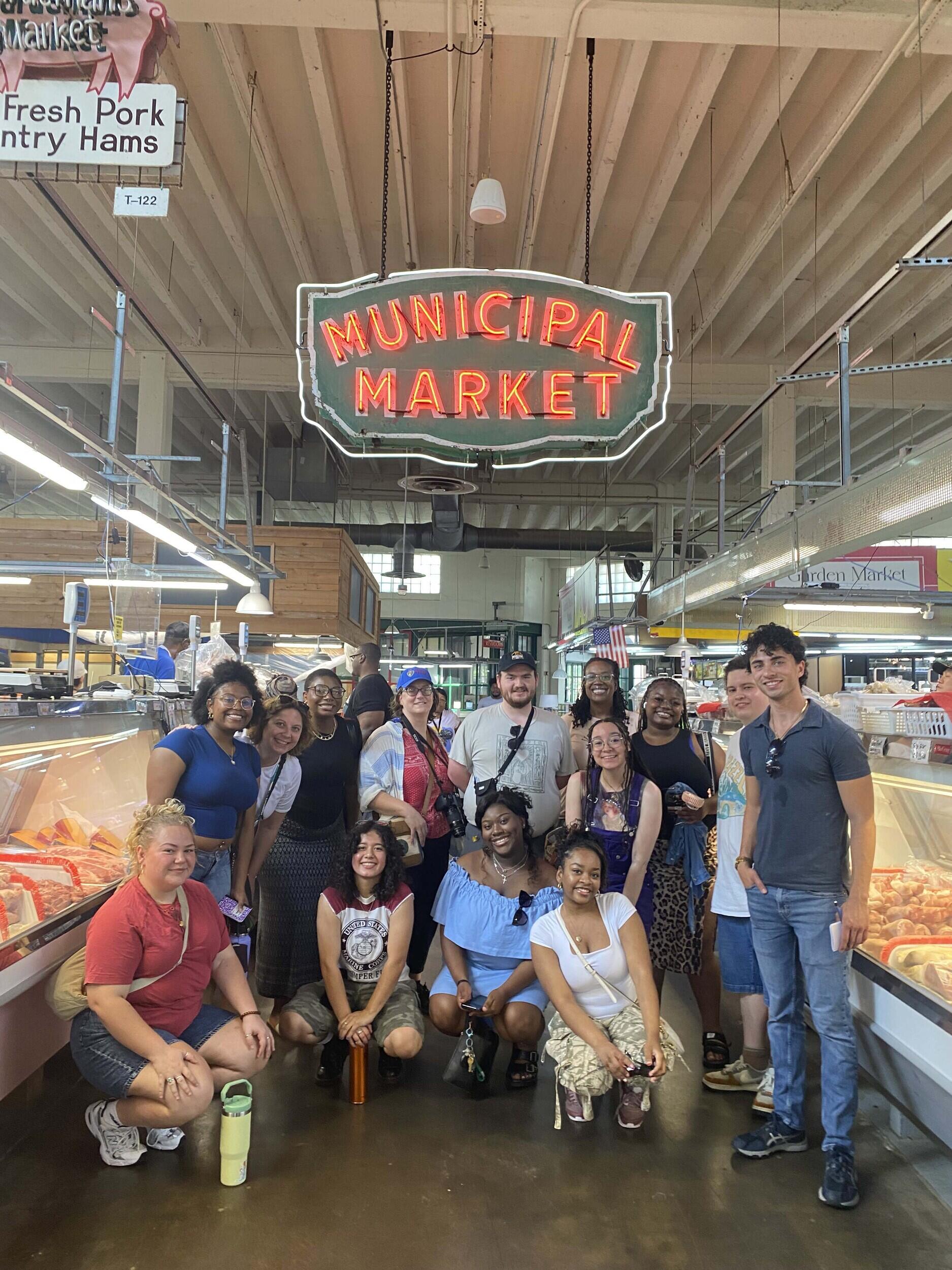 A photo of 13 people standing under a neon sign that says \"MUNICIPAL MARKET.\"