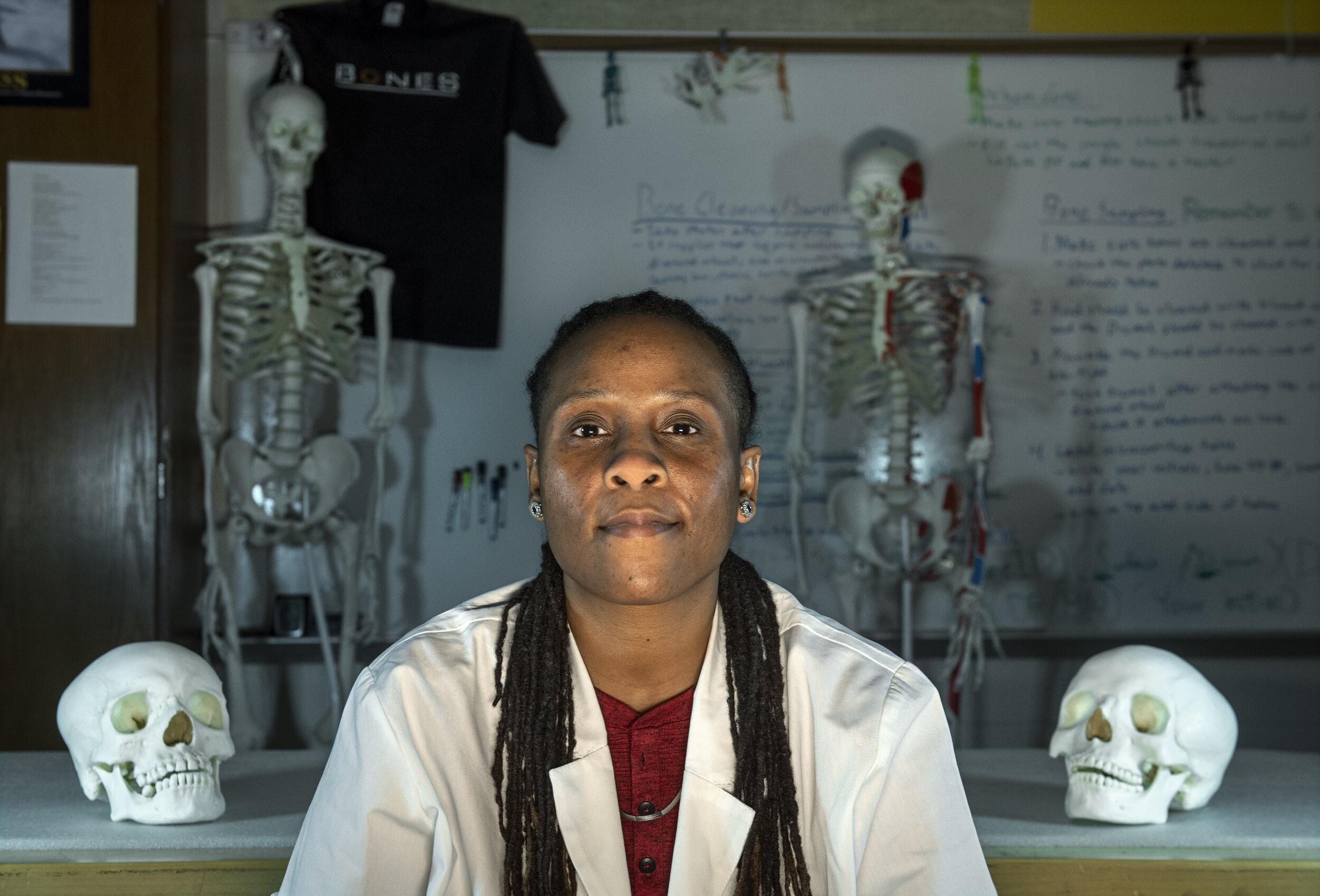 A woman wearing a lab coat sitting in between two skulls and two skeletons. Behind her is a white board with text written all over it. 