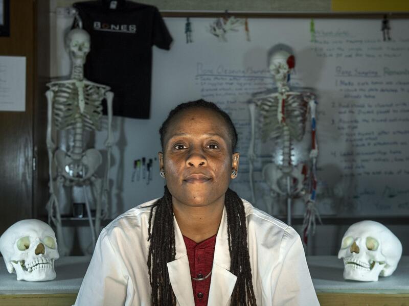A woman wearing a lab coat sitting in between two skulls and two skeletons. Behind her is a white board with text written all over it. 