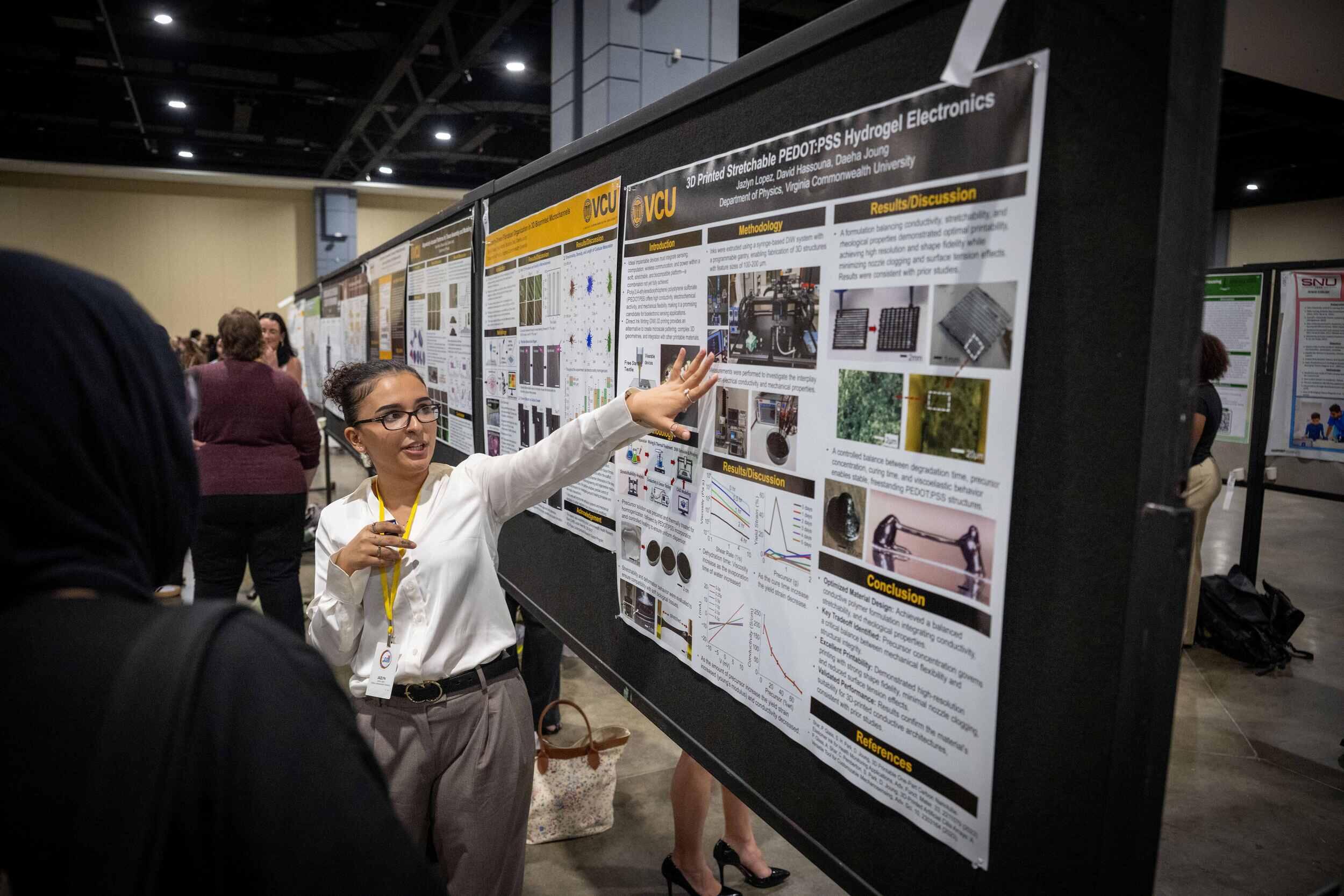 A woman in a white top and glasses points toward a research poster board as she speaks.