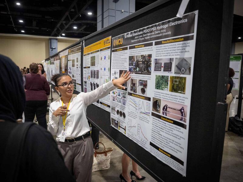 A woman in a white top and glasses points toward a research poster board as she speaks.