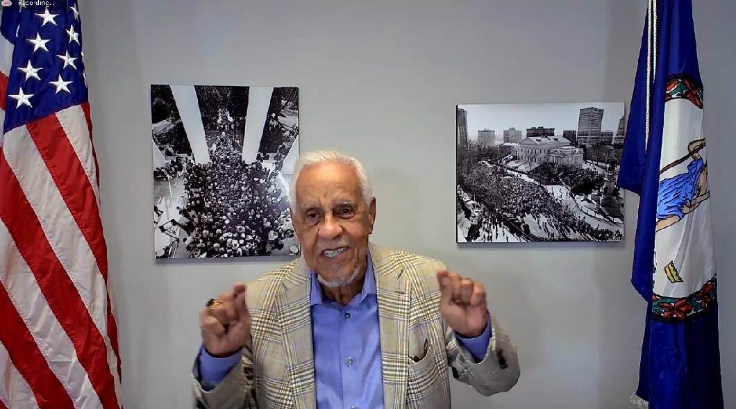 L. Douglas Wilder emphasizing a point with a gesture, sitting between an American flag and a Virginia flag.