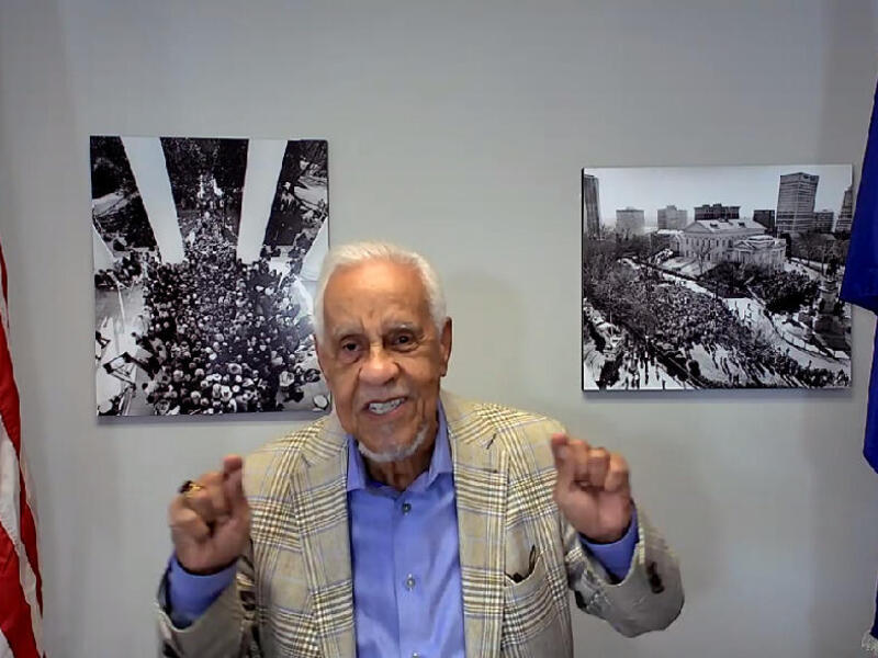 L. Douglas Wilder emphasizing a point with a gesture, sitting between an American flag and a Virginia flag.