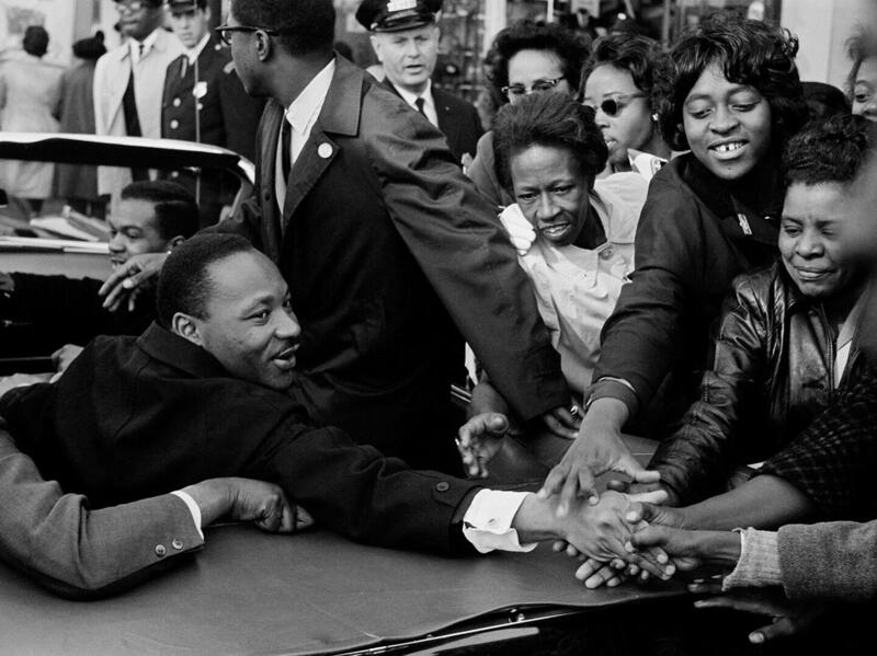 Martin Luther King Jr. reaches from a convertible toward onlookers with outstretched hands.
