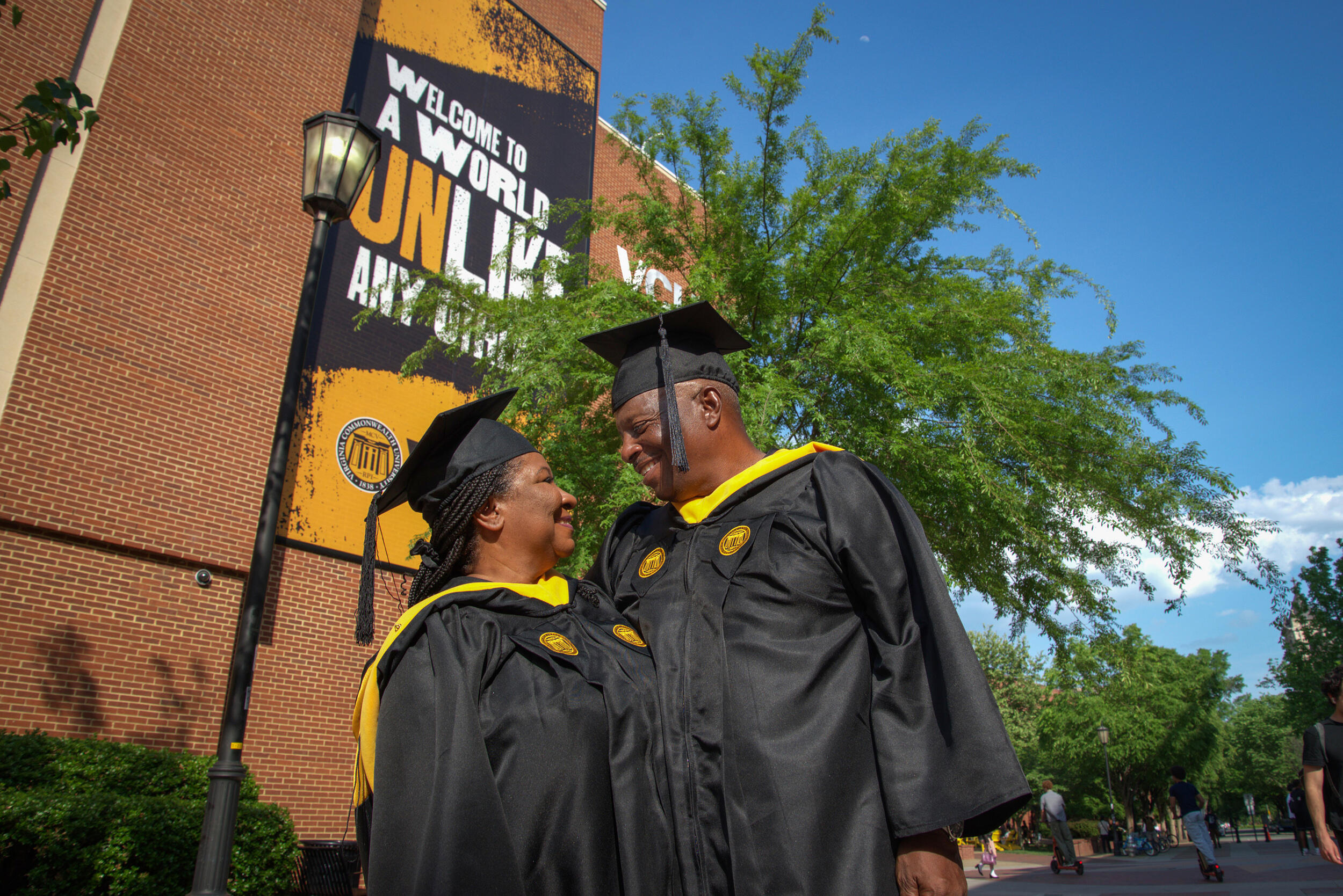 A photo of a man and a woman looking at each other lovingly. They are both wearing graduation cap and gowns. 
