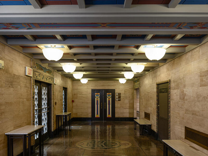 A photo of a hallway with lights and colorful tiles on the ceiling. The hallway has three doors on the left side and one on the right. At the end of the hallway is a doorway. On either side of the hallway are two benches. 