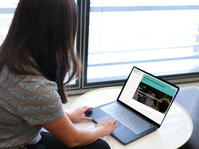 A photo of a woman from behind. She is sitting at a small table with a laptop on top of it. 