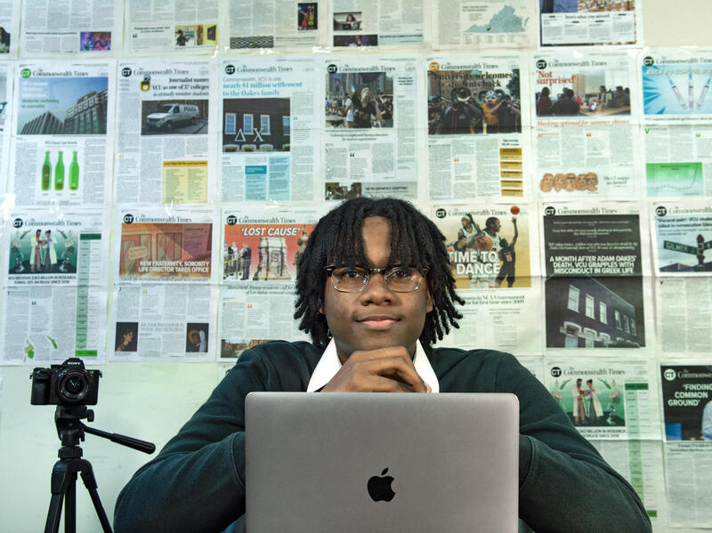 A man sitting in front of an Apple laptop with his hands under his chin. Behind him are layouts of newspaper pages. 